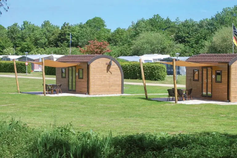 Zwei SeeLodges im Sonnenlicht auf der grünen Rasenfläche des Campingplatzes mit Holzwänden, einem runden Dach, 2 kleinen Fenstern und eine doppelflügelige Glastür, davor jeweills eine gepflasterte Terrasse mit Tisch, vier Stühlen und Sonnensegel im Süsel Seeparx
