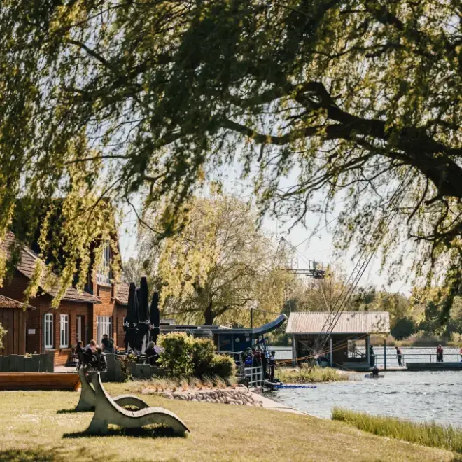 Blick über die gemütlichen Holzliegen auf der Liegewiese in Süsel Seeparx mit Blick rüber zum Hauptgebäude und auf die Wakeboard Park Seilbahn