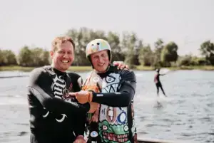 Christian Rumpel im Neoprenanzug mit einem Wakeboarder Arm in Arm am Ufer von Süsel Seeparx